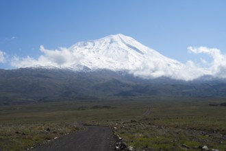 A snow-covered mountain rises majestically under a clear blue sky with scattered clouds, near
