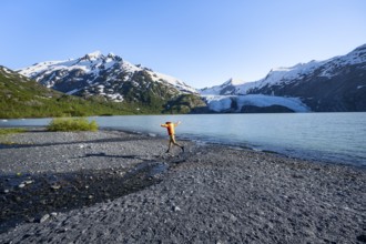 Young man jumping on a pebble beach on a glacial lake, Snowy Mountains and Portage Glacier on