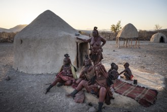 Married Himba woman with their babies in their arms sitting in front of the first woman's hut, in