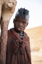 Himba girl leaning at a traditional clay hut, in the morning, traditional Himba village, Kaokoveld,