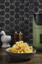 A bowl of crispy golden potato fries prepared in an air fryer is on a wooden kitchen table. Behind