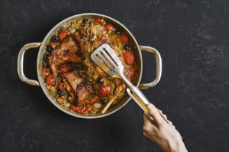 A hand uses a kitchen tongs to lift a tender rabbit leg from a pan filled with roasted tomatoes and