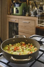 Chopped leeks and carrots sizzle in a large pot on a stovetop. The kitchen features wooden