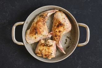 Two succulent rabbit hind legs and a saddle are cooked in a skillet. They are seasoned with herbs