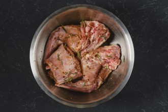 Two raw hind leg and saddle of rabbit is marinating with herbs and spices in a metal bowl. It is