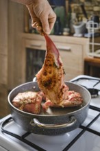 A person holds a marinated rabbit hind leg above a pot with other meat cooking inside. The kitchen