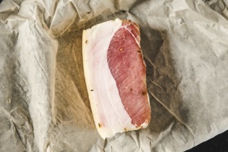 A slice of cured meat lies on a piece of parchment paper in a kitchen. The meat features a mix of
