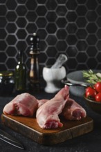 Rabbit hind legs sit on a wooden cutting board, surrounded by herbs, tomatoes, and kitchen tools.