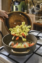 Sliced leeks and carrots are being poured from a wooden bowl into a pan on the stove. The warm