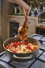 A hand holds a seasoned rabbit leg above a pot filled with fresh vegetables, including tomatoes and
