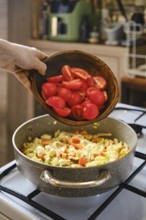 A person is adding chopped red tomatoes from a wooden bowl into a pot that is already filled with