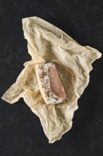 A rich piece of cured meat is neatly wrapped in crumpled brown paper resting on a dark surface.