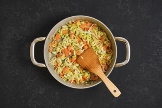 Chopped carrots and leeks are cooking in a large pan on a dark surface. A wooden spoon stirs the