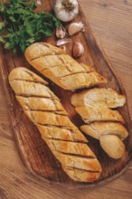 Garlic bread, baguette with garlic butter and herbs, on a wooden board, with herbs and garlic