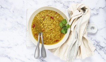 A bowl filled with hearty lentil soup sits on a marble surface, garnished with fresh herbs. A cloth