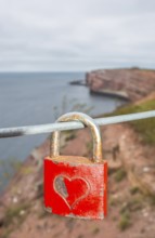Red padlock with engraved heart symbol on a wire rope in front of the steep coast, red cliff of