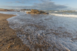 A wide sandy beach stretches out with scattered pebbles and beach shells. Gentle waves crash
