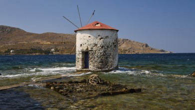 Historic windmill in the clear blue of the sea, surrounded by the rocky shore and bright sky, Agia