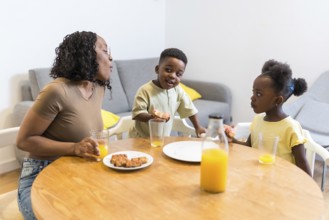 Mother and two children are sitting at a wooden table, eating breakfast and having orange juice