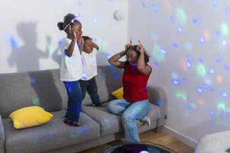 Mother making funny faces while kids dancing joyfully on the sofa, illuminated by colorful disco