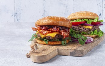 Two burgers with beef patty, fried bacon, cheddar cheese, tomatoes, on a light table