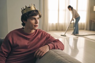 Portrait of teenage son wearing a golden crown while mother vacuums in background. Concept of
