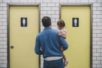 Father with young daughter hesitating in front of men's and women's restroom doors. Concept of