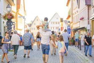 Families and pedestrians stroll through a lively old town street in sunny weather, 950 years of