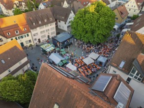 Aerial view of a lively town festival, surrounded by orange-coloured roofs and green trees in