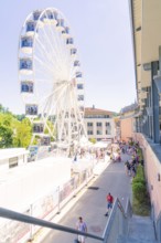 Ferris wheel next to a building with many people on a sunny promenade, Calw, Germany