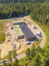 Building with solar roof in a construction site surrounded by forest, crane nearby, various