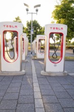 Several Tesla charging stations in an urban environment with sunlight and trees, Tesla Supercharger