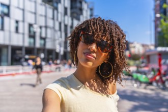 Portrait of a young woman with curly hair and sunglasses taking a selfie on a sunny day in the city