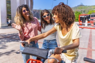 Three cheerful young women using smartphone and riding a sharing bike in a city center during