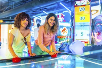 Two young women enjoying a friendly game of air hockey in a vibrant arcade, illuminated by colorful