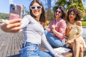 Group of female friends taking a selfie while enjoying pizza on a bench in a city park during a