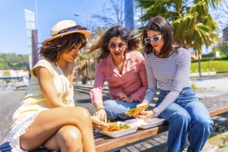 Three female friends enjoying a pizza lunch together on a park bench during a sunny summer day