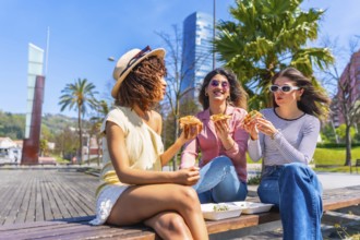 Three women savoring pizza slices while enjoying a sunny day on a park bench, sharing laughter and
