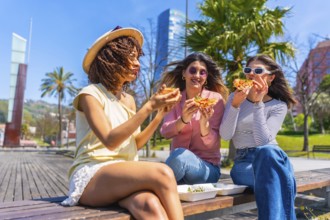 Three young women savoring delicious pizza while sitting on a bench in a vibrant city park,
