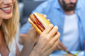 Woman enjoying a hot dog with ketchup, sitting at a picnic table, with a blurred man in the