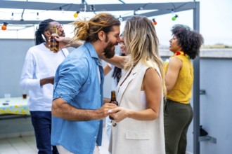 Happy couple dancing joyfully and sipping beer at a vibrant rooftop party, surrounded by diverse