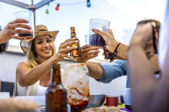 Cheerful friends raising glasses and bottles, making a toast during a rooftop party, celebrating