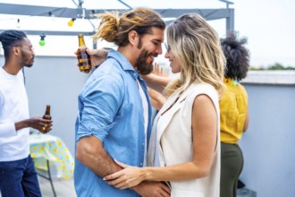 Happy couple embracing tenderly at a rooftop party, surrounded by friends enjoying drinks and good