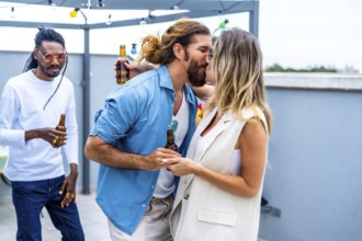 Young couple kissing and holding hands at rooftop party while friends drinking beer and having fun