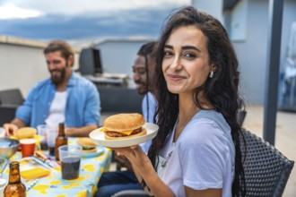 Young woman savoring a delicious burger while celebrating with friends at a lively rooftop barbecue