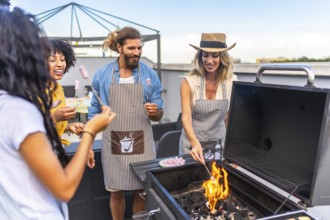 Happy friends wearing aprons are toasting marshmallows on a barbecue grill during a rooftop party