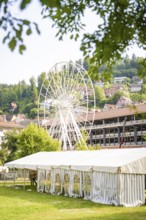 Ferris wheel behind a large white tent, surrounded by village houses and green hills, Ferris wheel