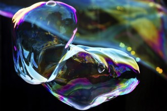 Irregular, iridescent soap bubble in the colours of the rainbow against a black background,