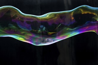 Large, iridescent soap bubble tube in the colours of the rainbow against a black background,