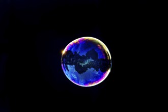 A shimmering and floating soap bubble in the colours of the rainbow against a black background,